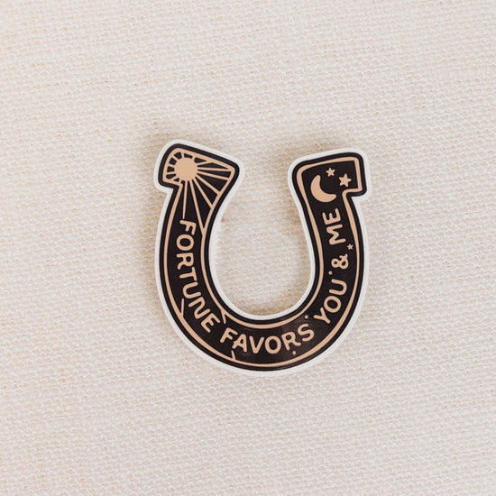 Horseshoe-shaped pin with 'Fortune Favors You & Me' text on a beige background