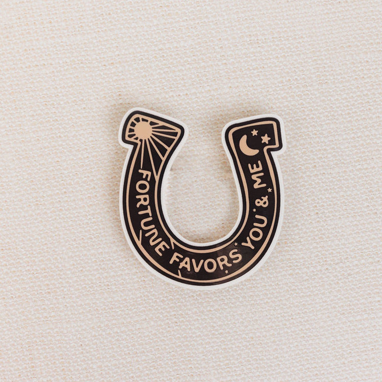 Horseshoe-shaped pin with 'Fortune Favors You & Me' text on a beige background